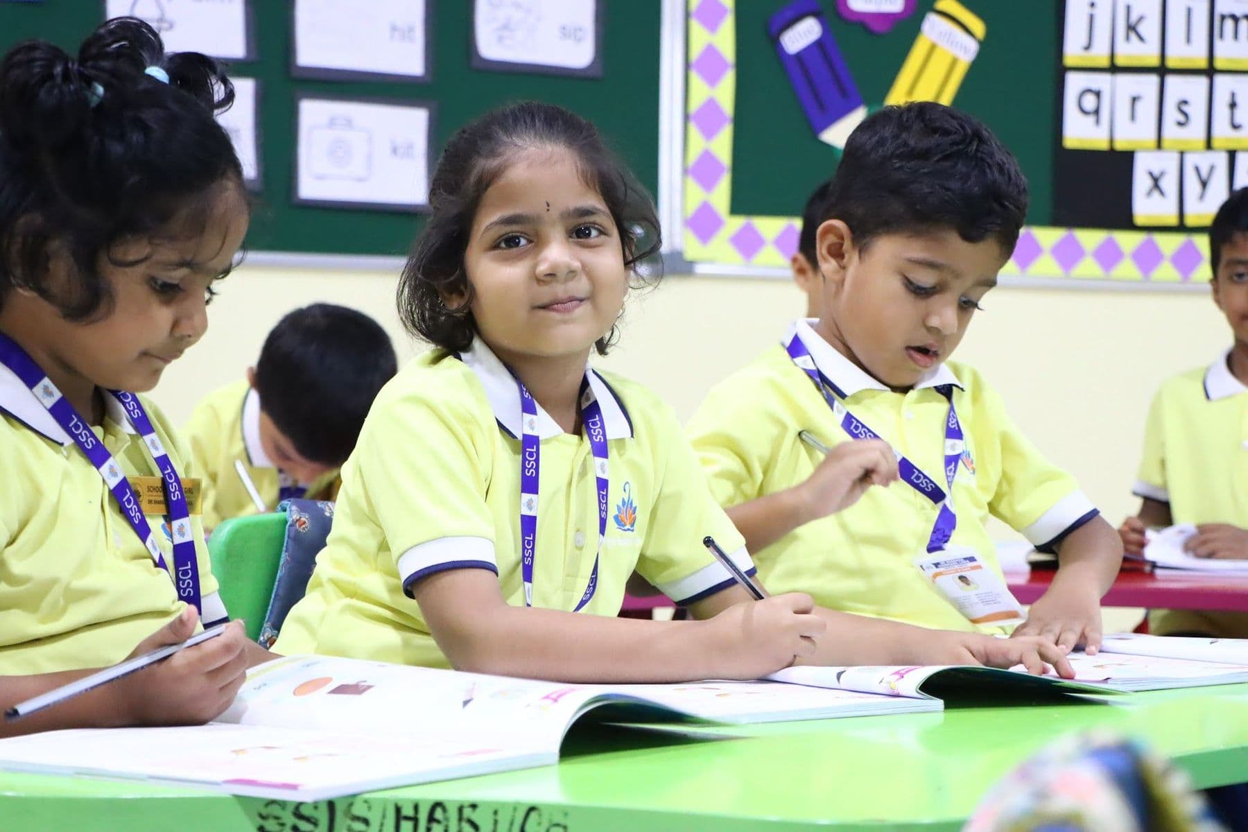 A smiling girl at her desk with classmates writing beside her