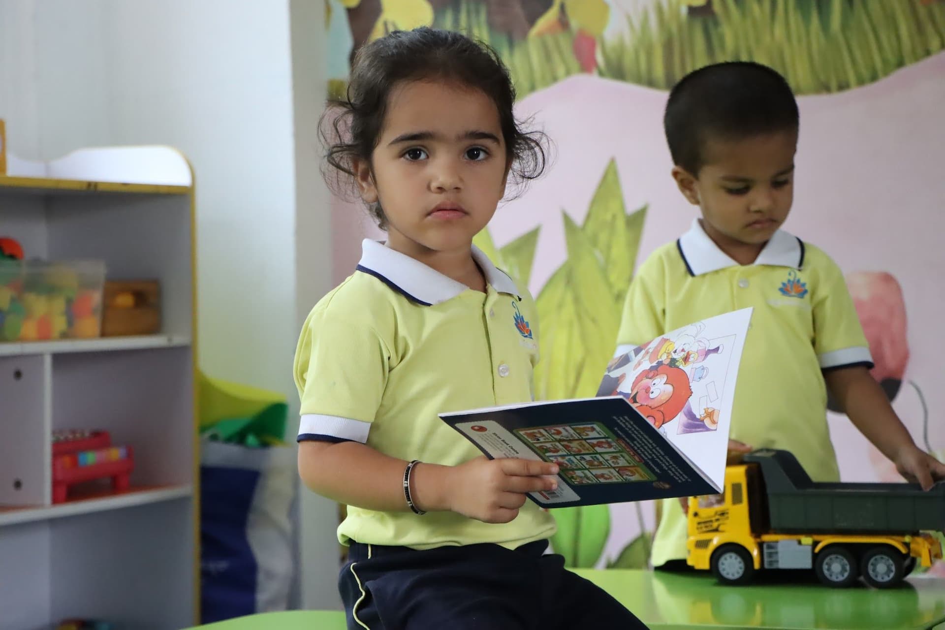 A girl reading while her friend plays with a toy truck