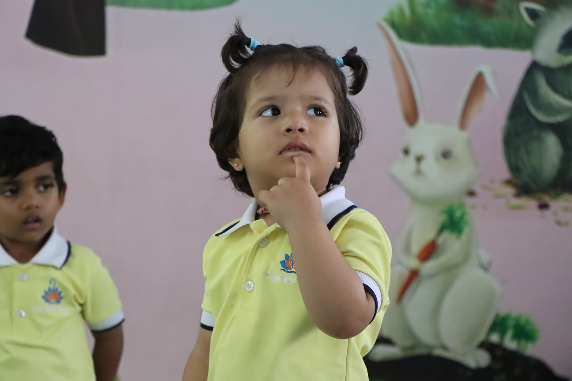 A toddler in a yellow uniform wondering, finger on her chin